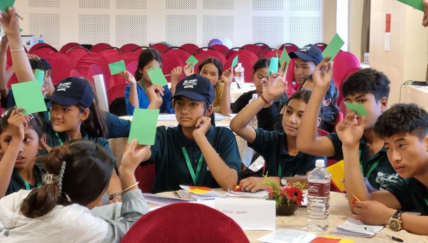 12 nuorta istuu pöytien ääressä. Kaikki ovat nostaneet yhden käden pystyyn ja näyttävät vihreää paperilappua.