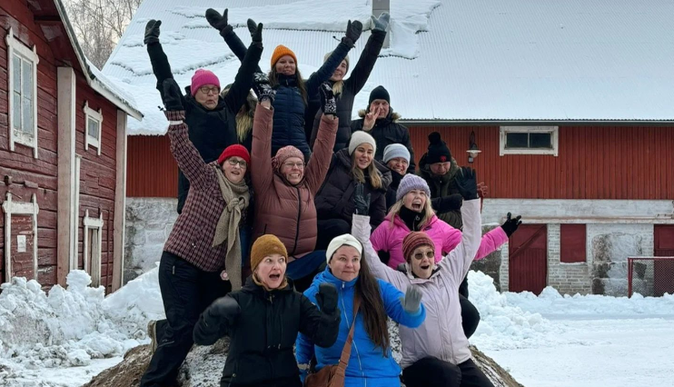 13 aikuista ulkona talvella punaisten matalien rakennusten välisellä pihamaalla. Ryhmä seisoo talvivaatteissaan tiiviisti yhdessä hiekkakasan päällä. Kaikki ovat nostaneet kätensä ilmaan ja hymyilevät leveästi tai huutavat.