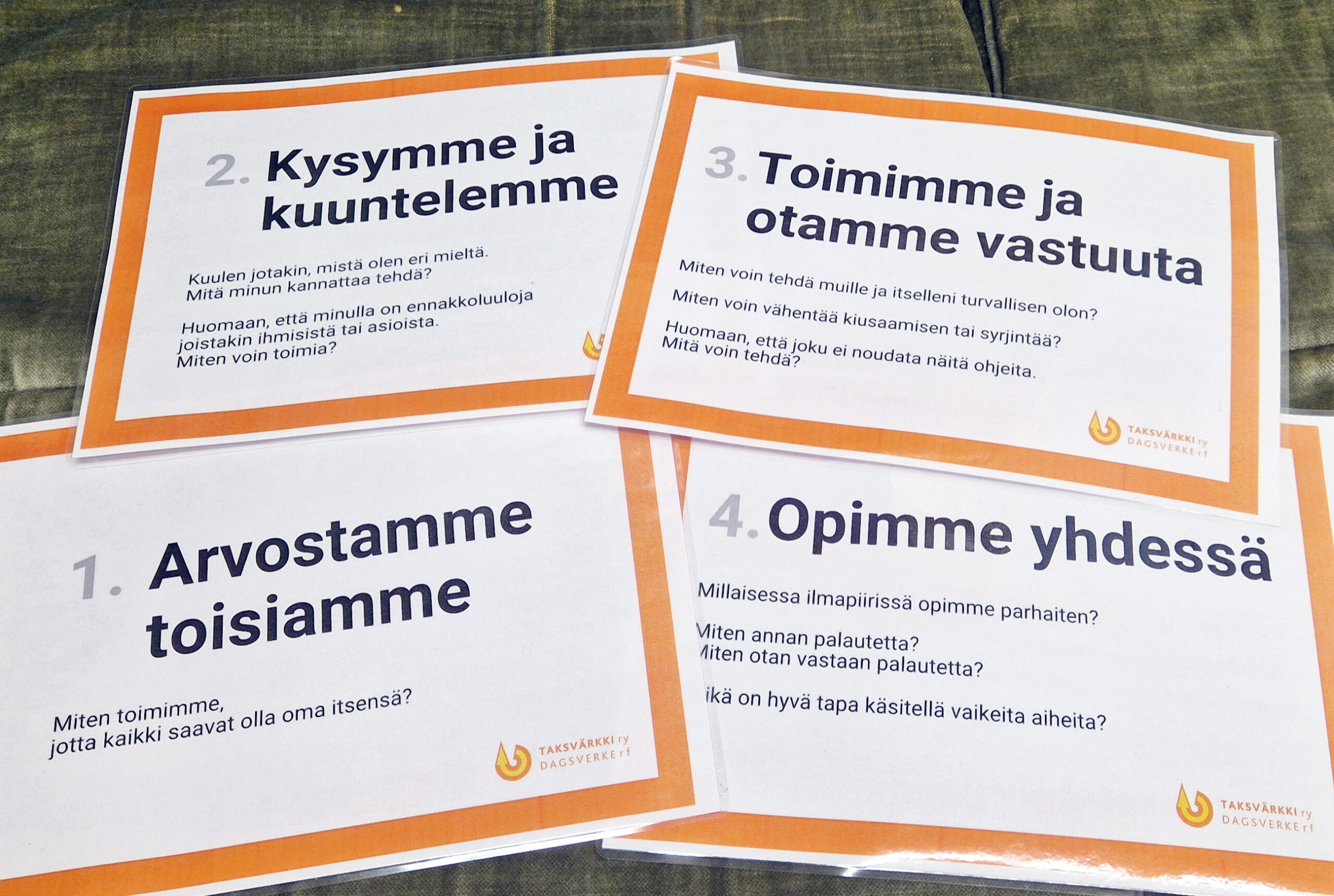 Kuvassa on neljä valkoista A4 paperin kokoista kylttiä, joissa on oranssit reunukset ja mustaa tekstiä. Kylttien otsikot ovat: 1. Arvostamme toisiamme, 2. Kysymme ja kuuntelemme, 3. Toimimme ja otamme vastuuta, 4. Opimme yhdessä.